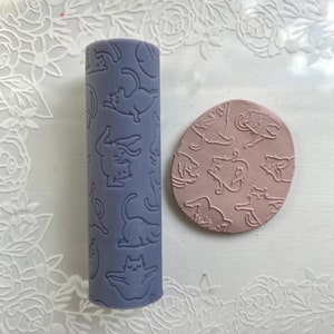 Peut inclure: Un rouleau à pâtisserie violet avec un motif répété de chats dans différentes poses. Le rouleau à pâtisserie est à côté d'un disque d'argile rose avec le même motif de chat.