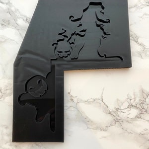 May include: Black Halloween-themed acrylic stencil featuring a witch silhouette holding a jack-o'-lantern. The stencil has a unique L-shape with a ghost cutout on the side. Ideal for crafting and seasonal decorations.
