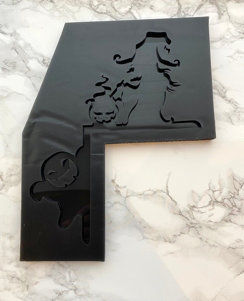May include: Black Halloween-themed acrylic stencil featuring a witch silhouette holding a jack-o'-lantern. The stencil has a unique L-shape with a ghost cutout on the side. Ideal for crafting and seasonal decorations.