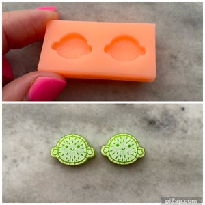May include: Two green and white resin lime-shaped earrings with a smiling face and arms. The earrings are shown next to a silicone mold with two lime-shaped cavities.
