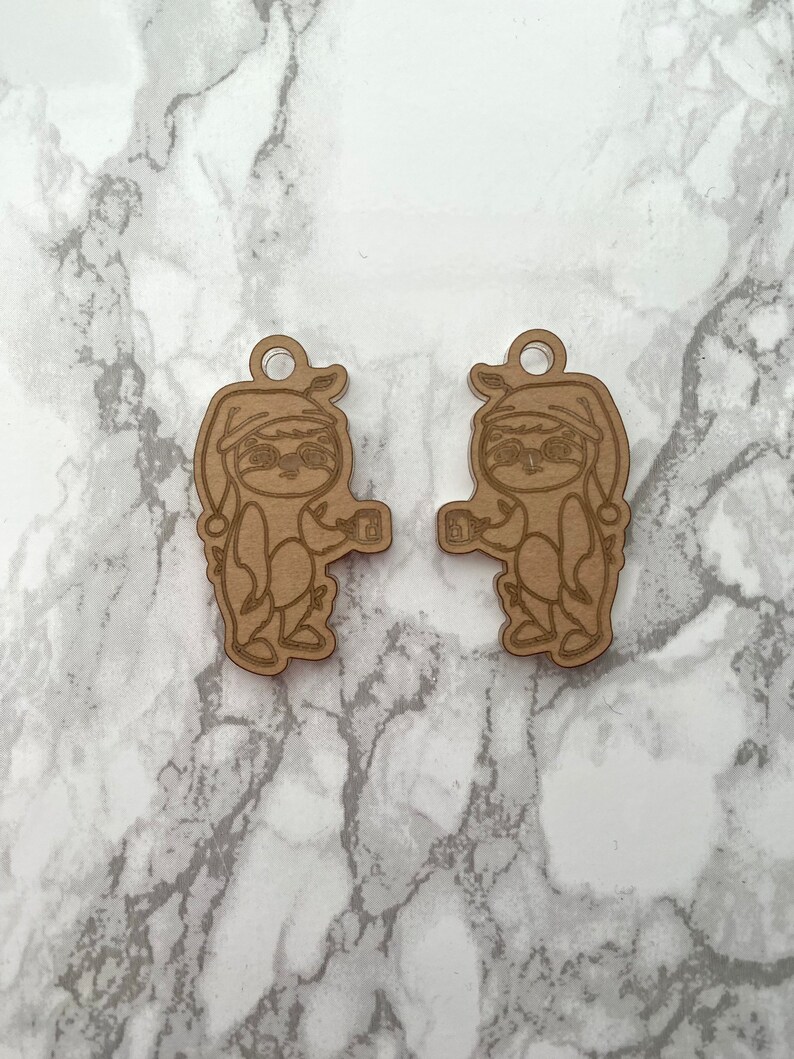 May include: Two brown wooden ornaments shaped like sloths wearing Santa hats and holding mugs. The ornaments are laser cut and have a rustic look.