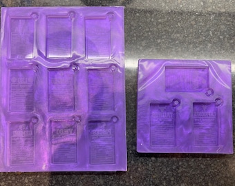 May include: Two purple silicone molds for making resin keychains. The larger mold has nine rectangular spaces, while the smaller mold has two rectangular spaces. The text "Traditional Tarot" is visible in the smaller mold.