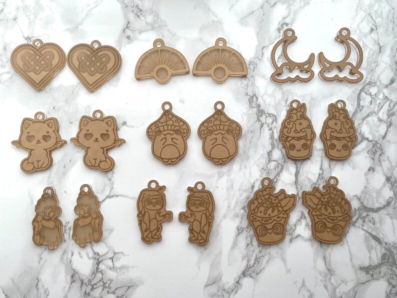 May include: A set of 12 laser cut wooden charms featuring various designs, including a cat, a mushroom, a cupcake, a sun, a moon, and a heart. The charms are made of natural brown wood and are perfect for adding a touch of whimsy to your crafts.