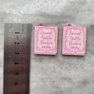 Puede incluir: Dos colgantes rosas con forma de libro con el texto "Dearest Gentle Readers". Los colgantes, de aproximadamente 1 cm de largo, tienen un dise&ntilde;o de borde floral. Est&aacute;n sobre un fondo gris claro.