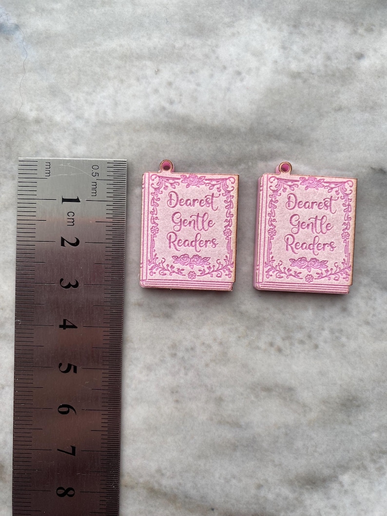 Puede incluir: Dos colgantes rosas con forma de libro con el texto "Dearest Gentle Readers". Los colgantes, de aproximadamente 1 cm de largo, tienen un dise&ntilde;o de borde floral. Est&aacute;n sobre un fondo gris claro.