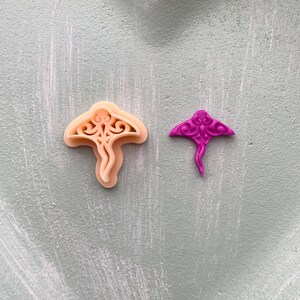 May include: Two cookie cutters in the shape of a manta ray. One is a light beige colour and the other is a bright purple colour.