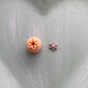 May include: Two snowflake-shaped cookie cutters on a light blue background. The larger cutter is a light peach colour and the smaller cutter is a light purple colour.