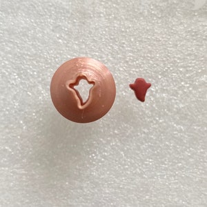 May include: A close-up of a pink plastic cookie press with a ghost-shaped cutout. A small, red, ghost-shaped cookie is next to the press.