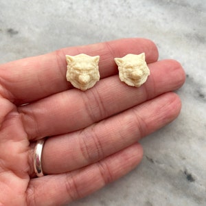 Puede incluir: Un par de pendientes de color crema con forma de cabeza de lobo. Los pendientes están hechos de un material liso y mate y tienen un diseño detallado de cabeza de lobo.