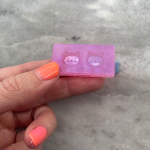 Puede incluir: Un molde de silicona rosa con dos diseños de cara de gato. El molde es translúcido y tiene una superficie texturizada.