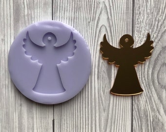 May include: A gold-colored angel-shaped ornament and a lavender silicone mold of an angel. The angel has outstretched wings and a circular head. The mold is round and has a detailed impression of the angel design.