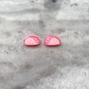 Puede incluir: Un par de pendientes de taco rosa. Los pendientes son pequeños y tienen un diseño sencillo.