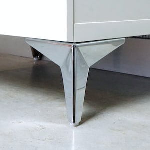 Op de afbeelding: Close-up van een wit kastje met chromen poten. De poten zijn driehoekig van vorm en hebben een glanzende afwerking.