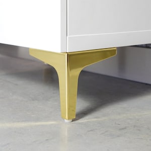 Op de afbeelding: Close-up van een wit kastje met gouden metalen poten. De poten zijn schuin en hebben een strak, modern ontwerp.