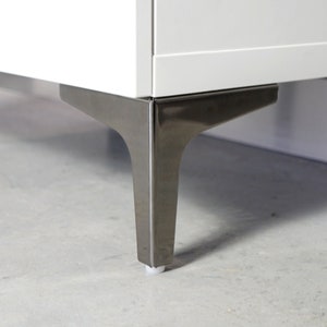 Può includere: Primo piano di un mobile bianco con gambe in metallo spazzolato. Le gambe sono angolate e hanno un design moderno. Il mobile è posto su una superficie grigio chiaro.