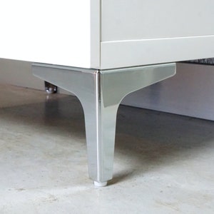 Può includere: Primo piano di un mobile bianco con gambe cromate. Le gambe sono angolate e hanno una finitura lucida.