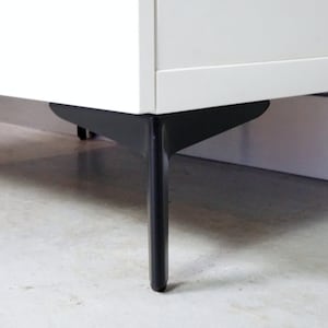 Op de afbeelding: Een witte kast met zwarte metalen poten. De poten zijn schuin en hebben een driehoekige vorm.