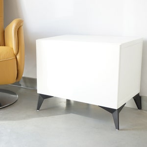 Op de afbeelding: Een moderne witte bijzettafel met zwarte, schuine poten. Op de achtergrond een gele leren draaistoel. De meubels staan tegen een witte muur en een lichtgrijze vloer.