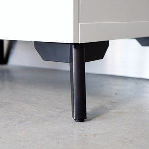 Könnte beinhalten: Ein weißer Schrank mit schwarzen Metallbeinen. Der Schrank hat ein modernes Design und ist aus hochwertigen Materialien gefertigt.