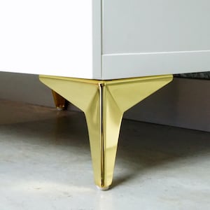 Op de afbeelding: Close-up van een wit kastje met gouden metalen poten. De poten zijn driehoekig van vorm en hebben een glanzende afwerking.