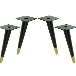 Op de afbeelding: Set van vier zwarte taps toelopende meubellepoten met gouden tips. Elke poot heeft een vierkante basis met een gat in het midden.