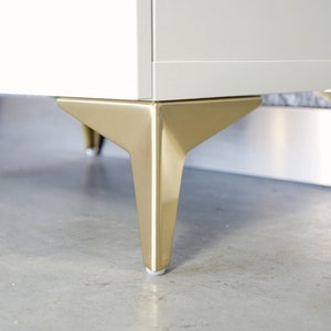 Puede incluir: Primer plano de un armario blanco con patas de metal dorado. Las patas son anguladas y tienen un acabado pulido.