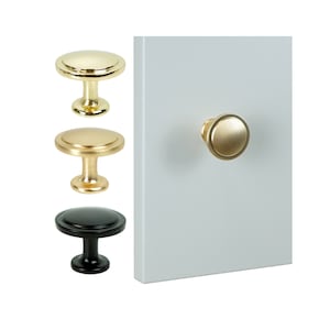 Könnte beinhalten: Drei Schrankknöpfe, einer schwarz, zwei goldfarben. Die Knöpfe sind rund mit einem erhöhten Rand und einem kleinen Stift zum Befestigen.