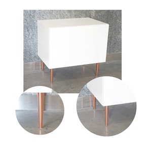 Może przedstawiać: Biała szafka z dwoma szufladami i miedzianymi nóżkami. Szafka stoi na betonowej podłodze.