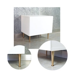 Op de afbeelding: Een witte kast met vier gouden metalen poten. De kast staat op een grijze betonnen vloer.