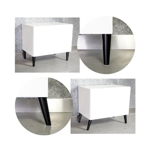 May include: A white bedside table with a minimalist design. The table has a rectangular shape and is supported by four black, tapered legs. The clean lines and contrasting colors create a modern aesthetic. The table is suitable for a bedroom or living room.