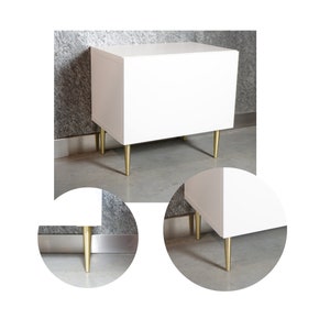 Könnte beinhalten: Ein weißer Schrank mit vier goldenen Metallbeinen. Der Schrank steht auf einem grauen Boden.