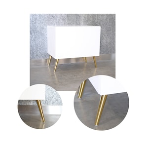 Könnte beinhalten: Ein weißer Schrank mit goldenen Metallbeinen. Der Schrank hat ein schlichtes Design und ist perfekt für ein modernes Zuhause.