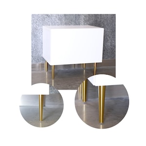 Op de afbeelding: Witte nachtkastje met gouden metalen poten. Het nachtkastje heeft een eenvoudig, modern ontwerp.