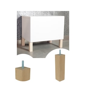 Può includere: Quattro gambe per mobili in legno con una vite in metallo filettata. Le gambe sono di colore marrone chiaro e hanno una forma quadrata. Le gambe sono fissate a un armadio bianco.