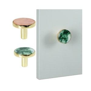 Könnte beinhalten: Zwei runde Schrankknöpfe mit goldenen Sockeln. Ein Knopf ist rosa mit weißen Wirbeln und der andere ist grün mit weißen Wirbeln.