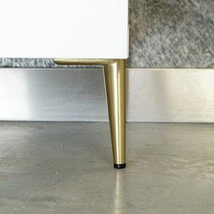 May include: Close-up of a white cabinet with gold metal legs. The legs are tapered and have a black rubber tip.