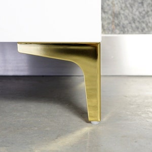 Può includere: Primo piano di una gamba di mobile in metallo dorato con una base bianca. La gamba è fissata a un armadio bianco.