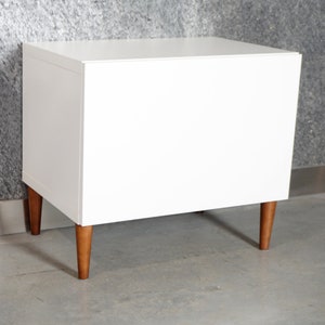 May include: White nightstand with wooden legs. The nightstand has a simple design and is made of a smooth, white material.