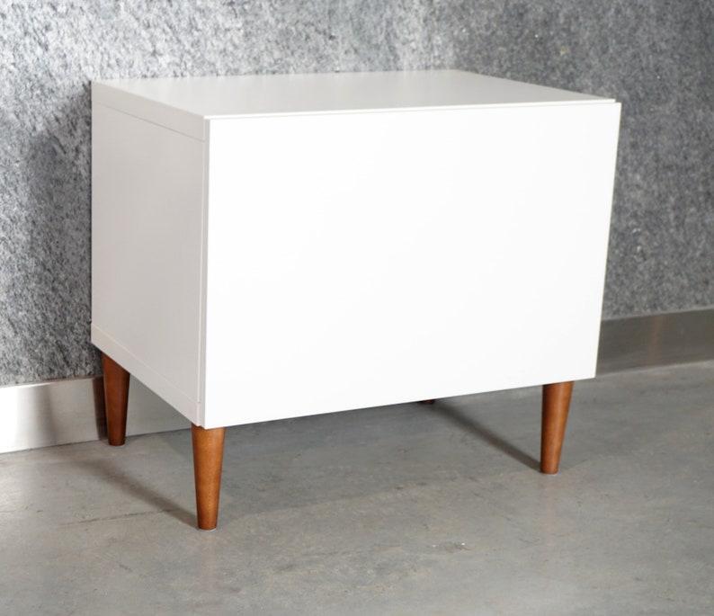 May include: White nightstand with wooden legs. The nightstand has a simple design and is made of a smooth, white material.
