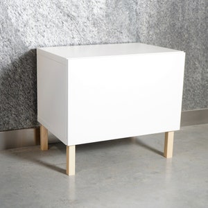 Puede incluir: Un armario de almacenamiento rectangular blanco con cuatro patas de madera. El armario tiene una superficie lisa y plana y está hecho de un material resistente.