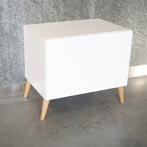 Puede incluir: Un armario blanco con patas de madera. El armario tiene un diseño sencillo y está hecho de un material blanco liso.