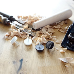Può includere: Primo piano di strumenti e materiali per la lavorazione del legno su una superficie in legno. Gli strumenti includono una pialla a mano, uno scalpello e un tassello di legno. Ci sono anche quattro piedini per mobili, due bianchi e due neri.