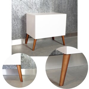 May include: White bedside table with a single drawer and four wooden legs. The table has a modern design with a white finish and natural wood legs.