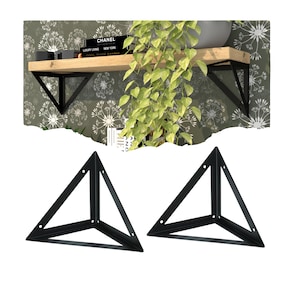 Puede incluir: Dos escuadras de estantería de metal negro con forma triangular, con agujeros pretaladrados para el montaje. Las escuadras se muestran sobre un fondo de una estantería de madera con libros y una planta, y un papel pintado con motivos.