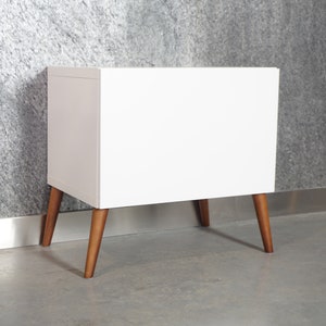 May include: White nightstand with a simple design and wooden legs. The nightstand has a smooth, flat surface and is perfect for a bedroom or living room.