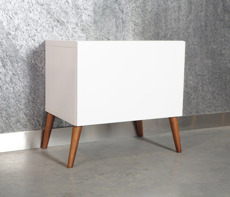 May include: White nightstand with a simple design and wooden legs. The nightstand has a smooth, flat surface and is perfect for a bedroom or living room.