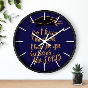 Op de afbeelding: Een ronde wandklok met een zwart frame. De marineblauwe wijzerplaat heeft witte wijzers en uurmarkeringen. Gouden tekst leest "For I know the plans I have for you declares the LORD" met een afstudeerhoed.