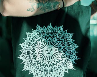 May include: Black tote bag featuring a detailed white mandala design. The intricate pattern is centered on the bag, creating a striking contrast against the dark fabric. The bag is held by a person, showcasing the design's prominence.
