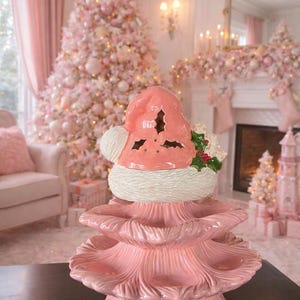 Peut inclure: Un sapin de Noël en céramique rose avec un chapeau de Père Noël. L'arbre a plusieurs niveaux avec des bords festonnés. Le chapeau de Père Noël a des détails découpés et est garni de fausse fourrure blanche. Du houx et des baies sont placés sur le chapeau. L'arrière-plan présente un sapin de Noël rose et des décorations.