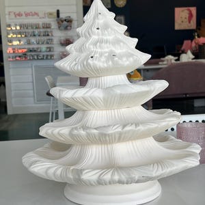 Könnte beinhalten: Ein weißer Weihnachtsbaum aus Keramik mit einem mehrstufigen Design. Der Baum hat eine strukturierte Oberfläche und mehrere kleine Löcher für Lichter. Der Sockel hat eine einfache, runde Form. Ein dekorativer Artikel für die Weihnachtszeit.